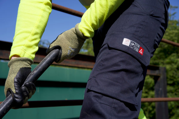 A person bends over showing navy blue PPE trousers and a fluorescent yellow overall and gloves while holding a metal pole. To support information about EN ISO 11611 Protective clothing for use in welding and allied processes