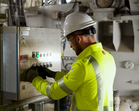 A man in a white helmet and fluorescent jacket has two hands on a large electrical box in a dimly lit industrial room. To support information about NFPA 2112 - Flame Resistant Clothing standards