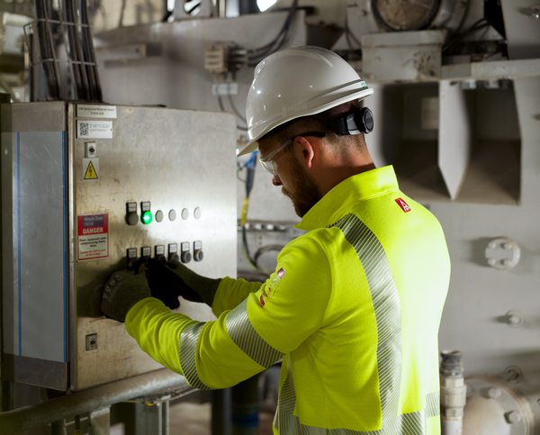 A man in a white helmet and fluorescent jacket has two hands on a large electrical box in a dimly lit industrial room. To support information about NFPA 2112 - Flame Resistant Clothing standards