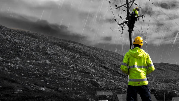 One man in a yellow helmet and fluorescent yellow jacket supervises another man wearing the same clothing who is suspended from a power cable working. Behind is a grey moorland background. To support information about ASTM F1891.