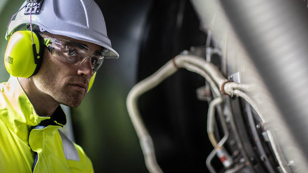 A man wearing a fluorescent yellow jacket, white helmet and fluorescent yellow ear defenders looks towards the camera. To support information about EN 352 - Hearing Protectors
