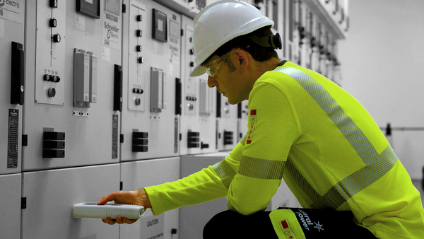 A man in a white helmet and flourescent yellow overall bends down to take a reading from a large grey electrical machine. To support information on EN ISO 11612, PPE Clothing to protect against heat and flame