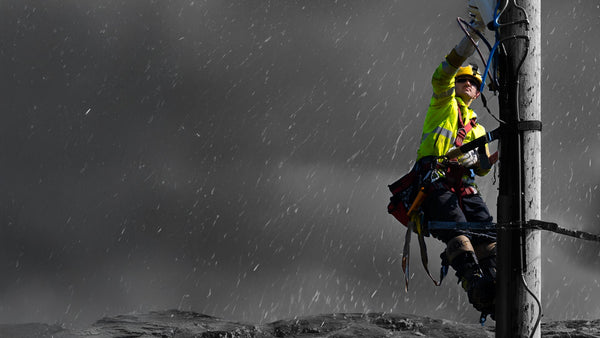 A man in a fluorescent overall and helmet repairs and electrical system while hanging from it. A grey background sits behind. To support information about EN 343 Protective clothing - Protection against rain