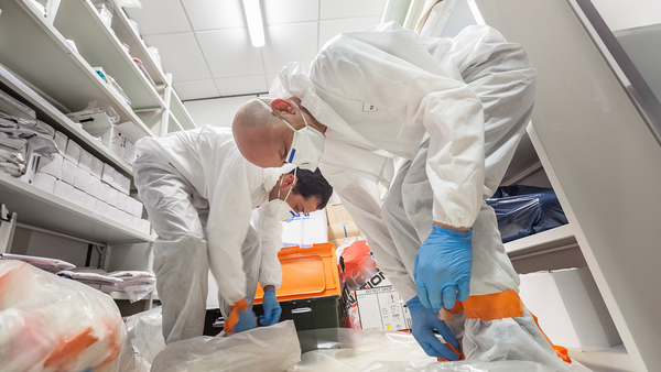 Taken from the floor two men in white protective suits and respirators bend down to work on the floor in a white industrial room to support information on EN ISO 13982 1