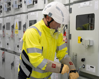 A man in a white helmet, gloves and fluorescent yellow jacket works on a large machine which occupies the rest of the picture. To support information on IEC 61482 - Protective clothing against the thermal hazards of an electric arc