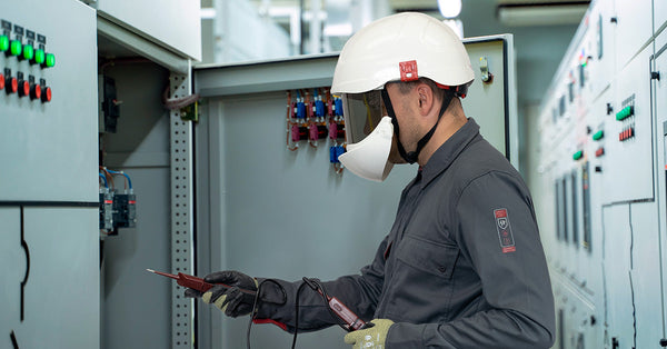 A man in a grey shirt with white helmet, visor and face mask looks into a metal cupboard while holding a device. Used to support information for EN 1149-5 Safety Standards on the Electrostatic Properties of Protective Clothing