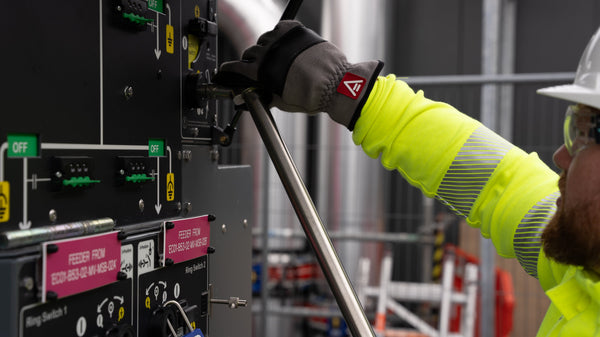 A man wearing a white helmet, fluorescent yellow jacket and gloves lifts a metal tube up to a machine to support information about EN 407 - protective gloves against thermal risks